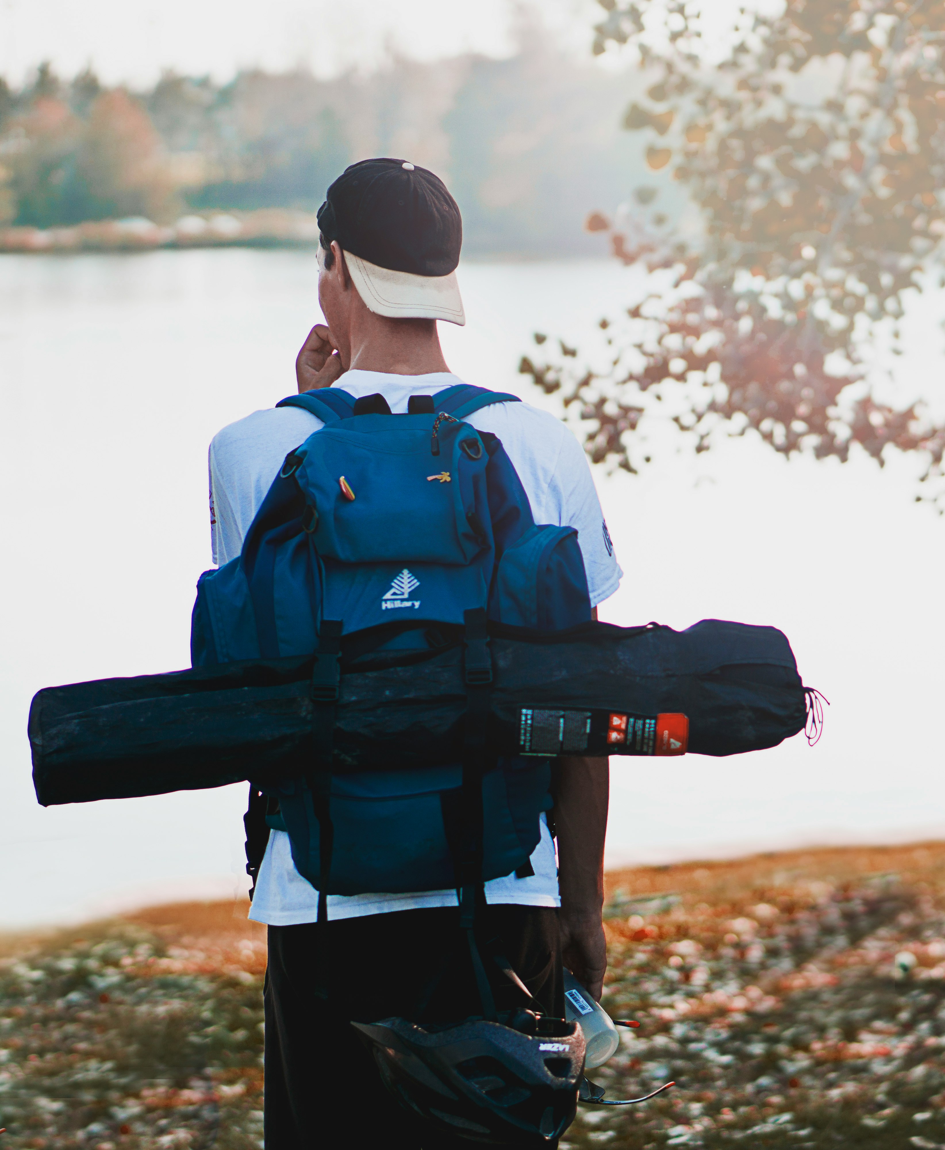 man wearing hiking backpack and tent bag standing near calm body of water